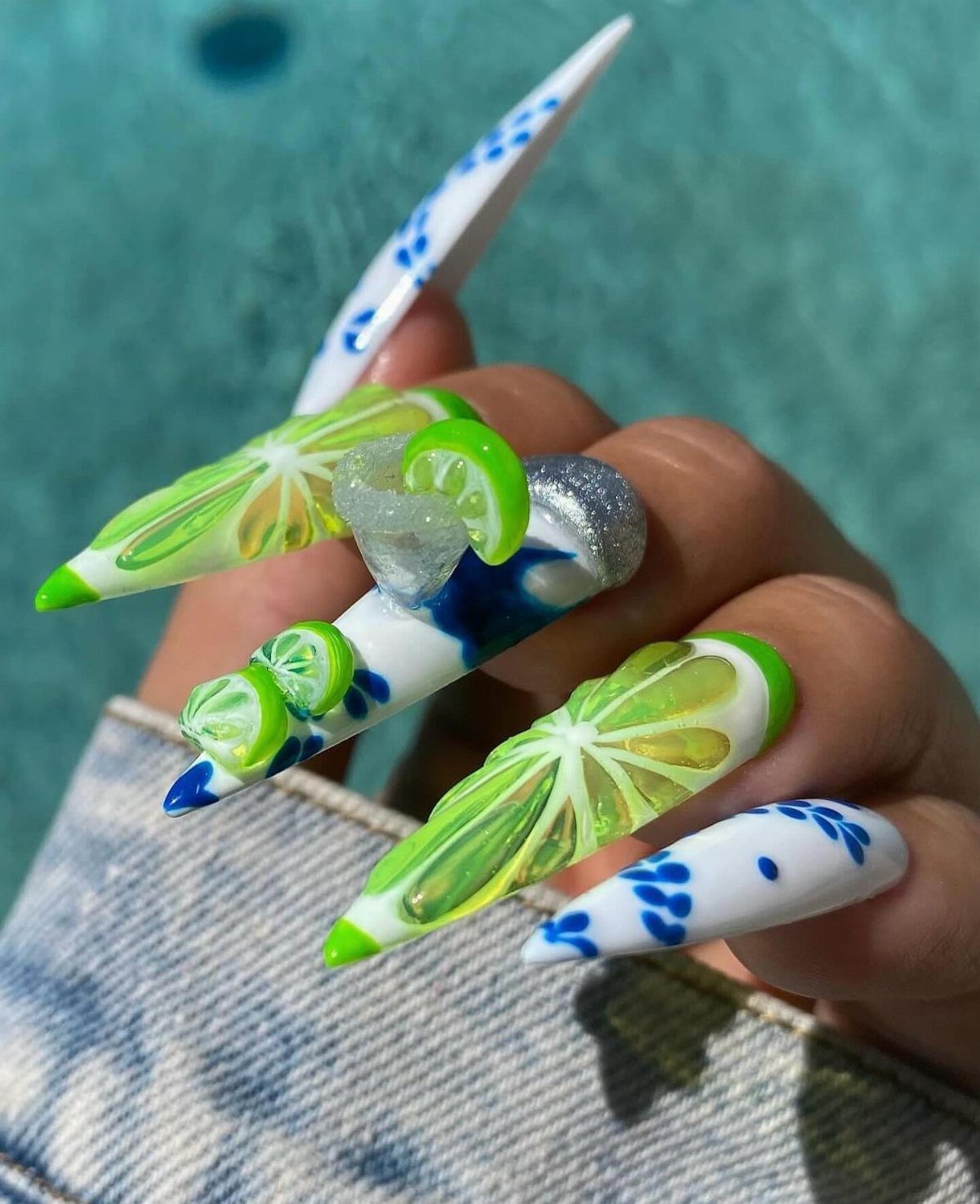 Long pointed nails with lime slice art and blue floral patterns by a pool
