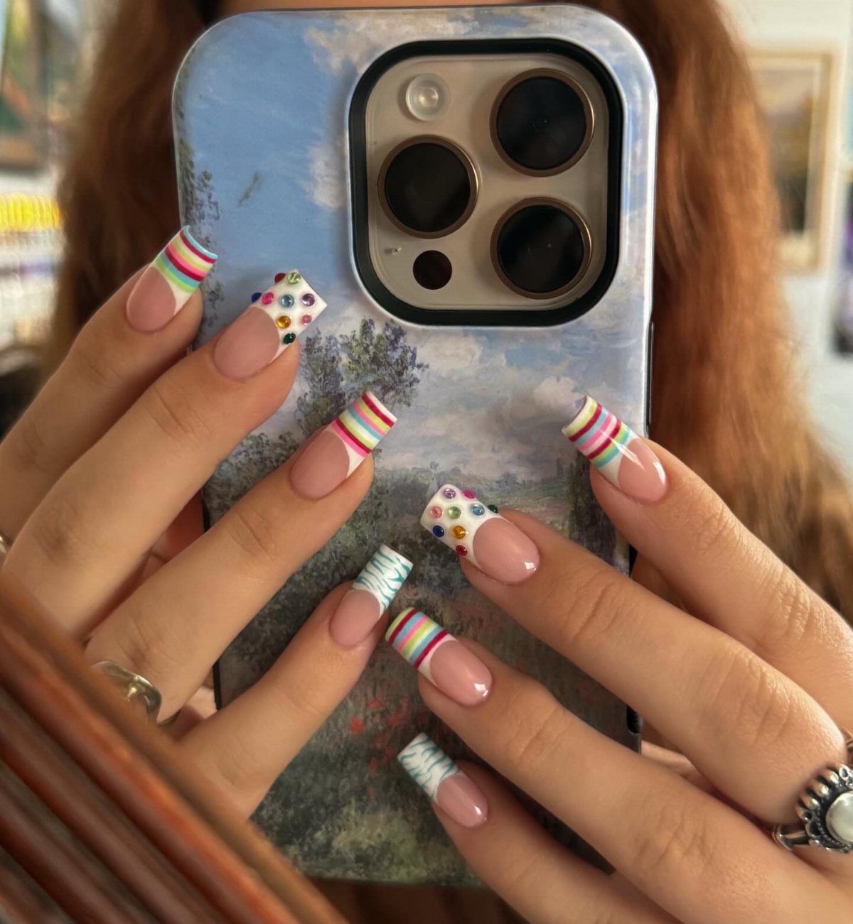 Manicured nails featuring colorful horizontal stripes and rhinestone accents holding a smartphone.