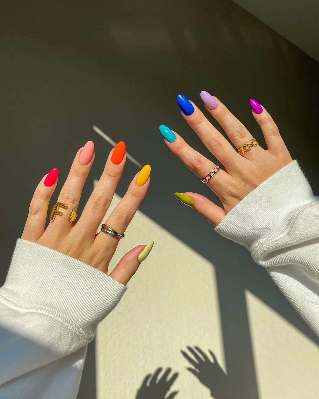 Rainbow colored almond shaped nails on two hands wearing gold and silver rings.