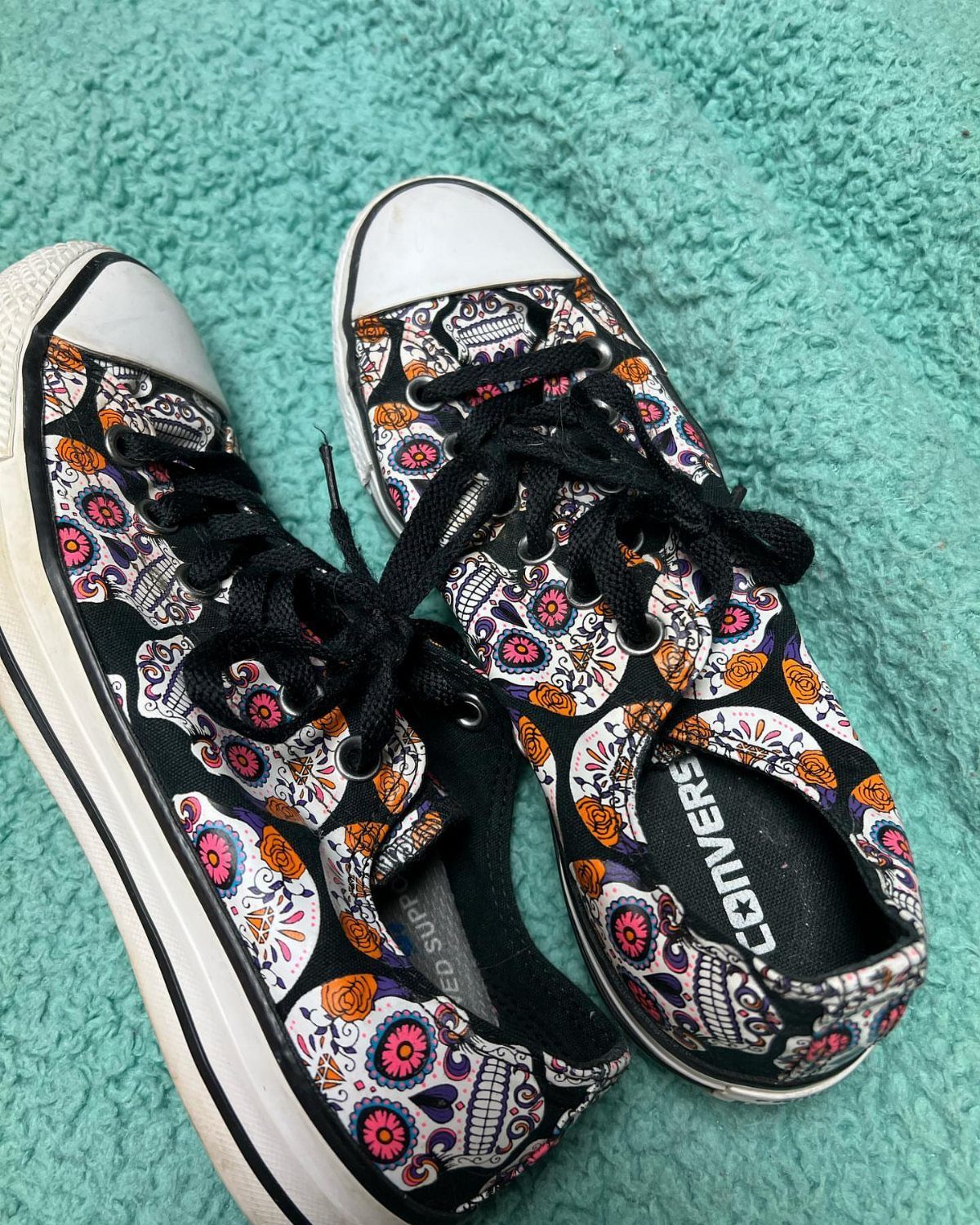 Black canvas sneakers with colorful sugar skull patterns on a teal carpet.