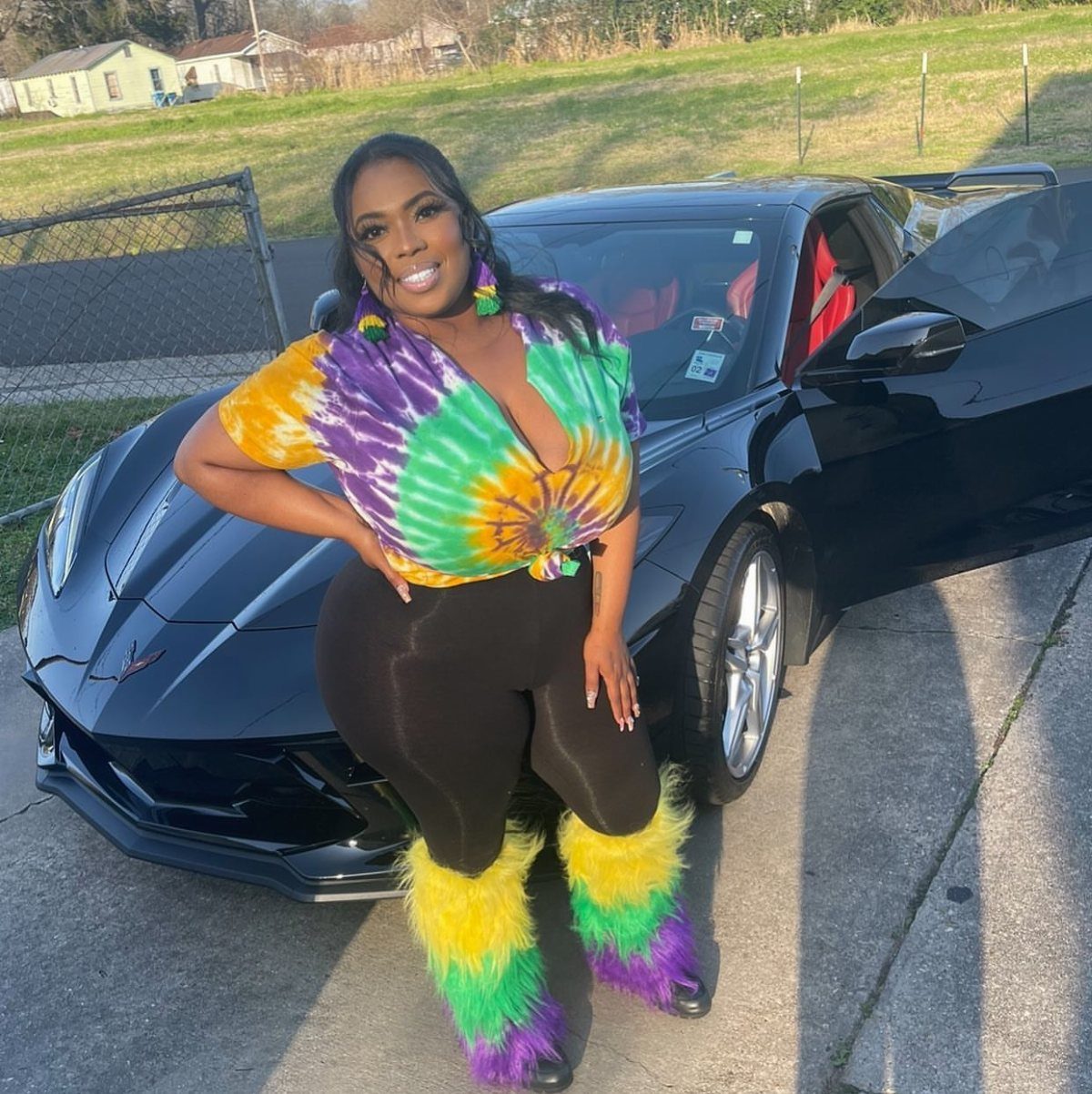 Woman in tie-dye top and colorful faux-fur leg warmers posing by black car