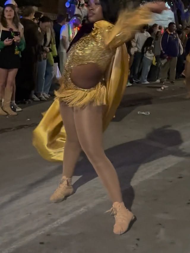 Person in a gold sequined fringe outfit and cape dancing on a street.