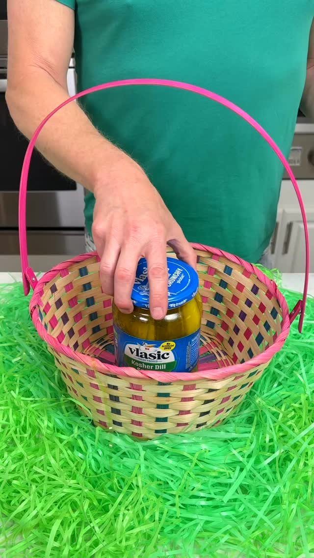 Hand placing a pickle jar into a woven Easter basket on green grass
