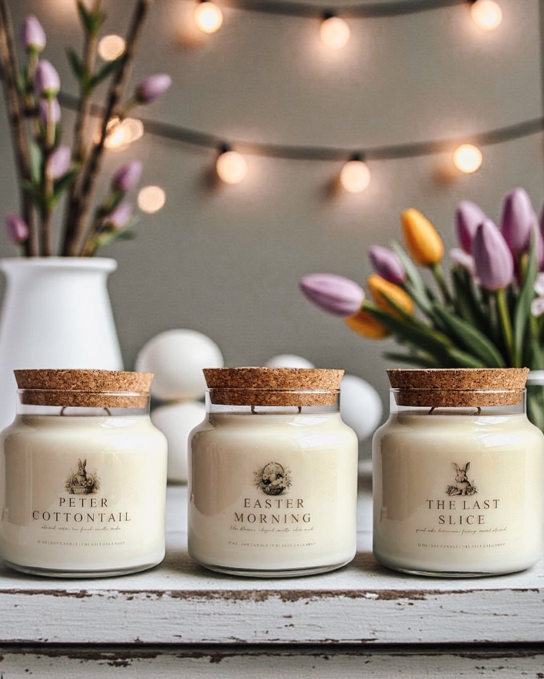 Three white scented candles with bunny labels on a rustic white wooden table.