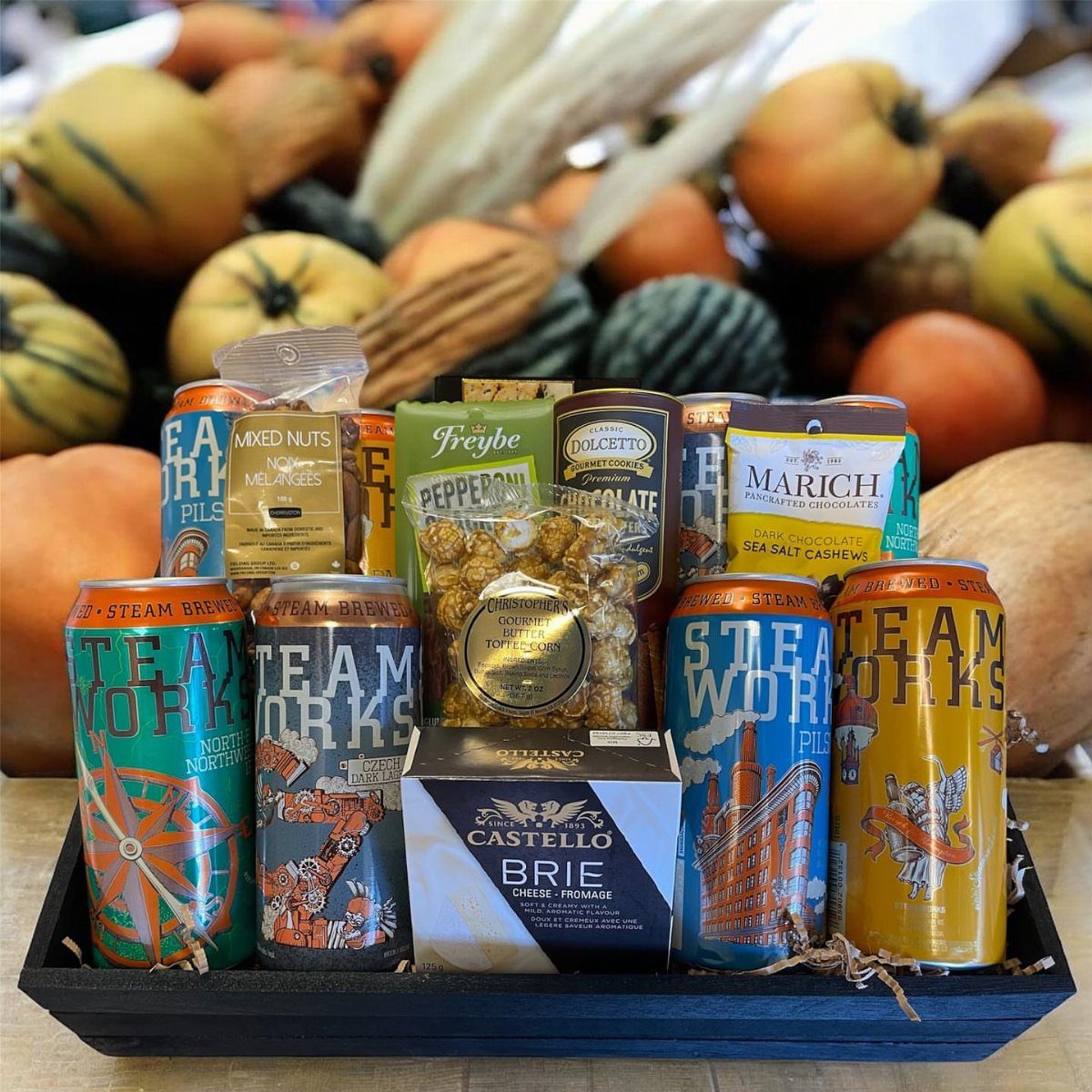 Craft beer basket with colorful cans, gourmet snacks, and brie cheese in a tray.