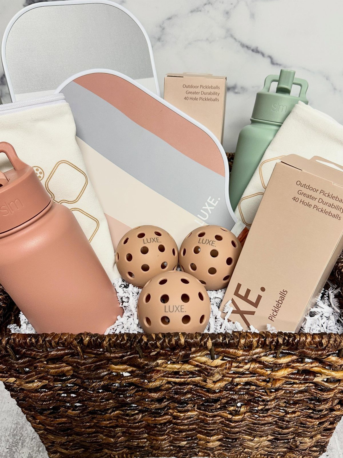 Pickleball paddle, balls, and water bottles arranged in a brown woven gift basket.