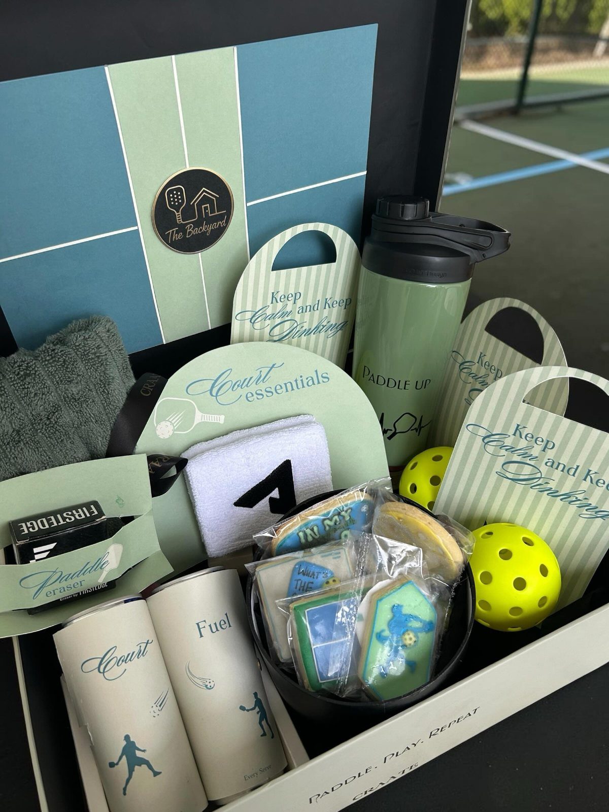 Gift box with tennis gear, water bottle, and cookies on a tennis court.