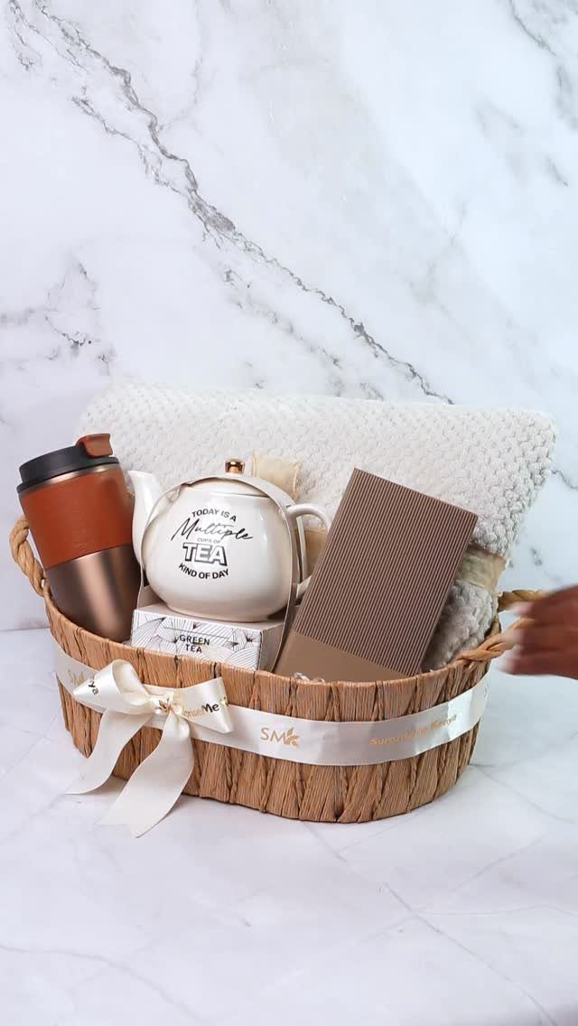 Woven gift basket with travel mug, teapot, notebook, and white blanket on marble.
