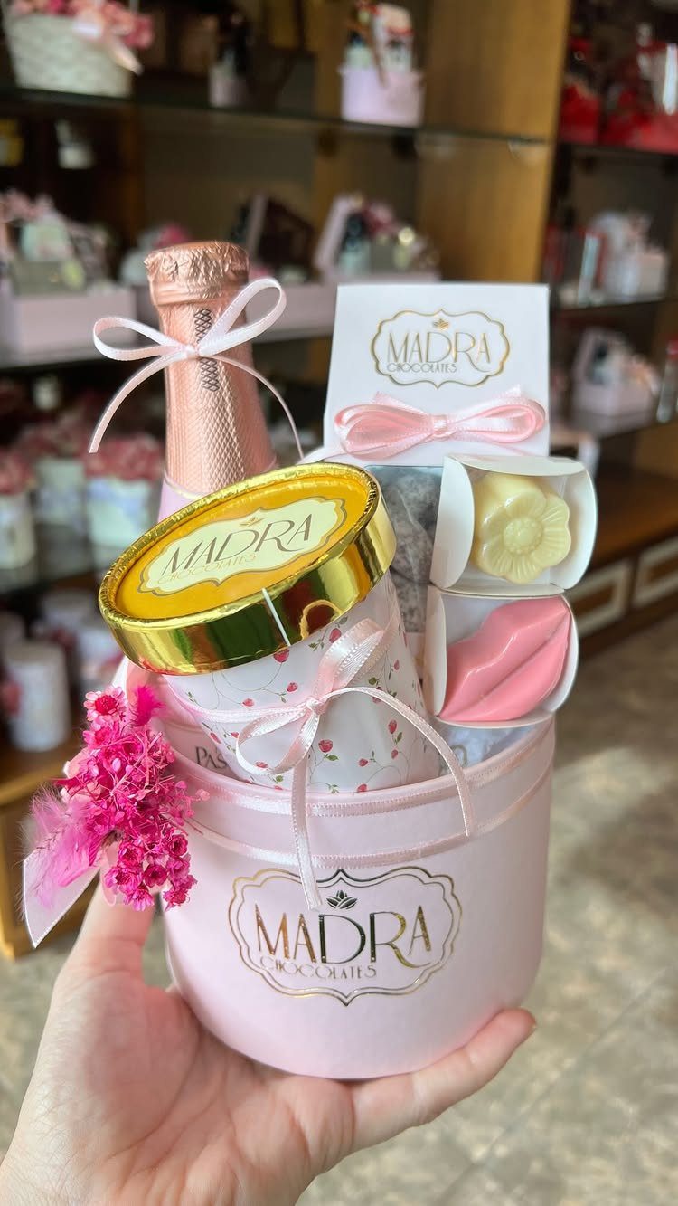 Pink gift basket with sparkling wine, chocolates, and flowers held in a hand.