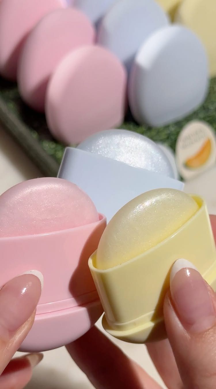 Hands holding open pink and yellow egg-shaped lip balm containers with pastel eggs.