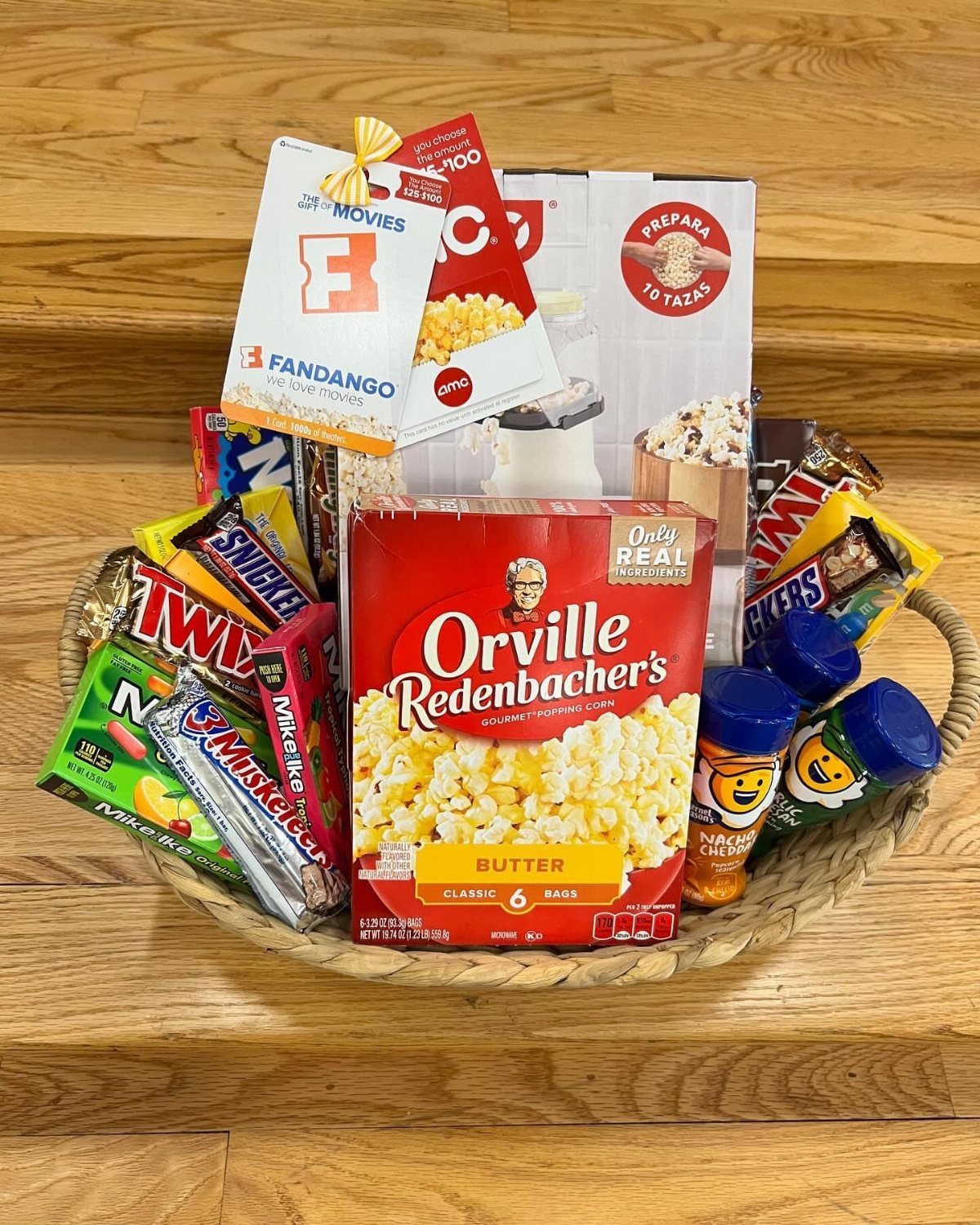 Woven basket filled with popcorn, movie tickets, and assorted candy on wooden floor.
