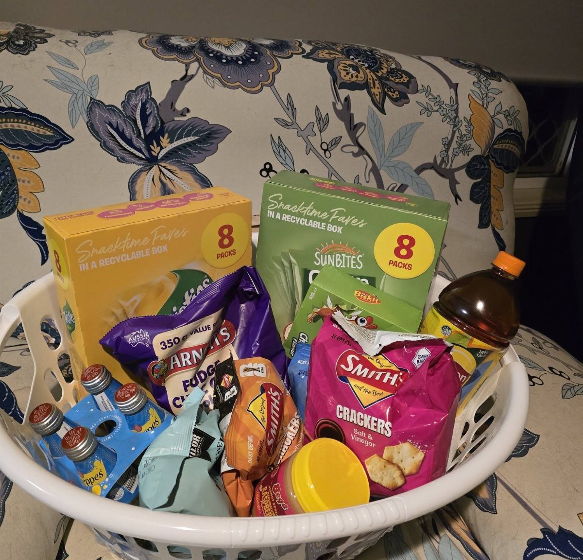 White plastic colander filled with assorted snack boxes, crackers, and bottled drinks.