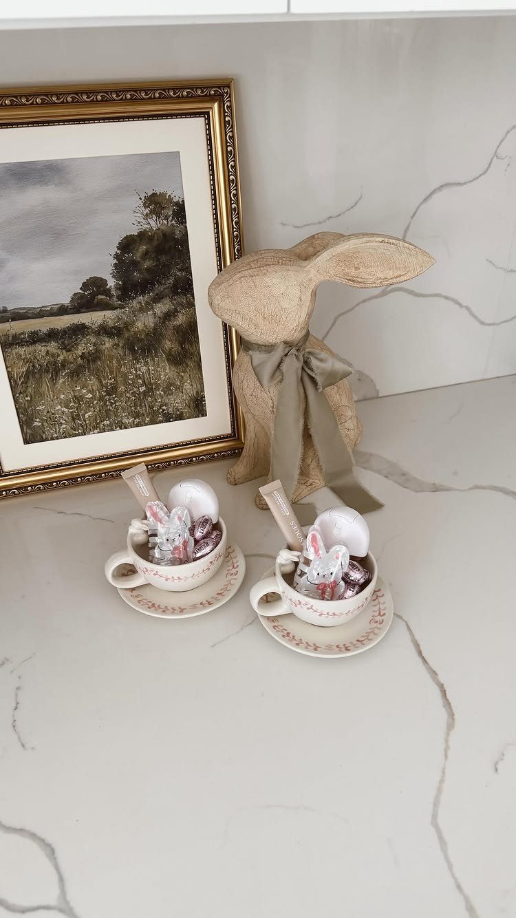 Two teacups with miniature Easter bunnies next to a straw bunny and framed art.