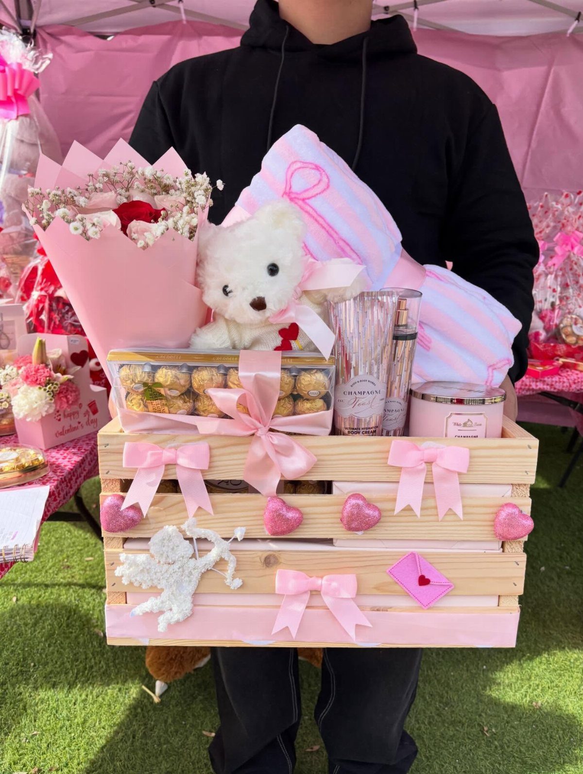 Gift crate with teddy bear, pink pillow, flowers, and chocolates held by person.