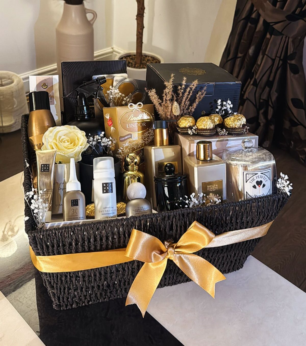 Black woven gift basket with gold ribbon containing perfumes, cosmetics, and chocolates.