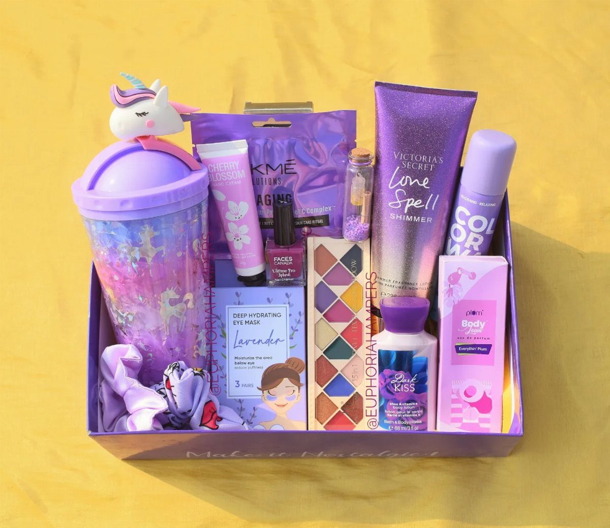 Purple gift box containing beauty products, a unicorn tumbler, and makeup on yellow background.