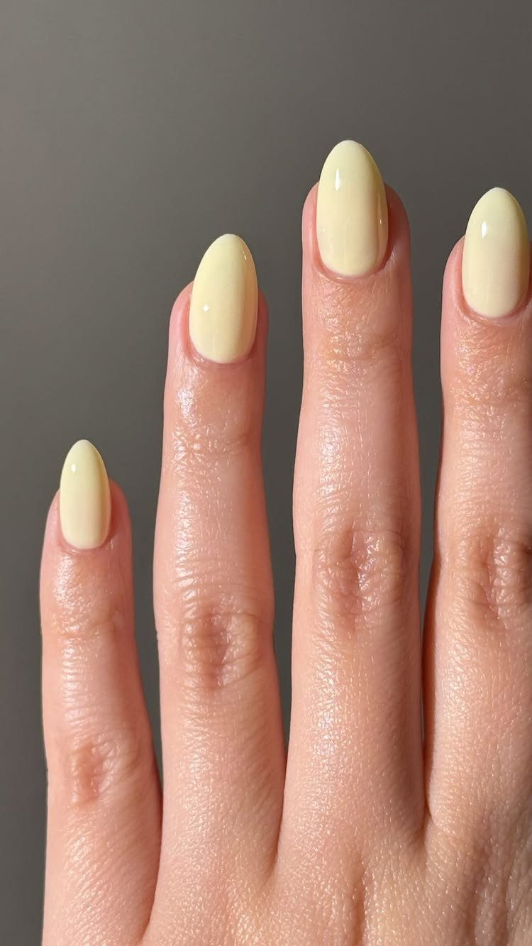 Pastel yellow nail polish on oval-shaped fingernails against a grey background