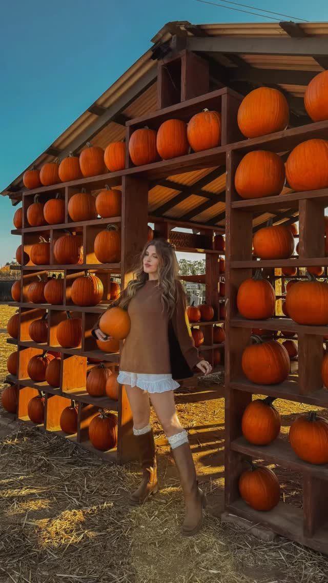 Woman in brown sweater and white mini skirt posing among orange pumpkins.