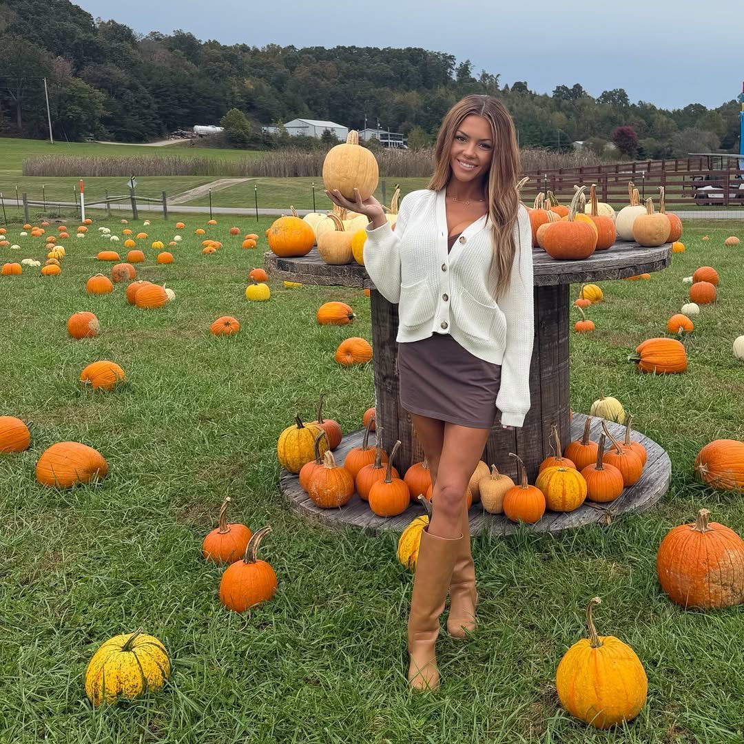 Woman in white cardigan and brown mini skirt holding a pumpkin in a field