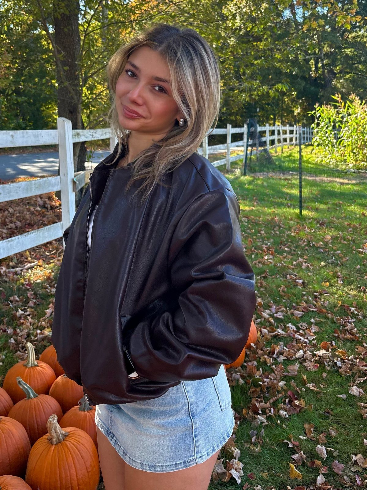 Woman in brown leather jacket and denim skirt standing by pumpkins and fence
