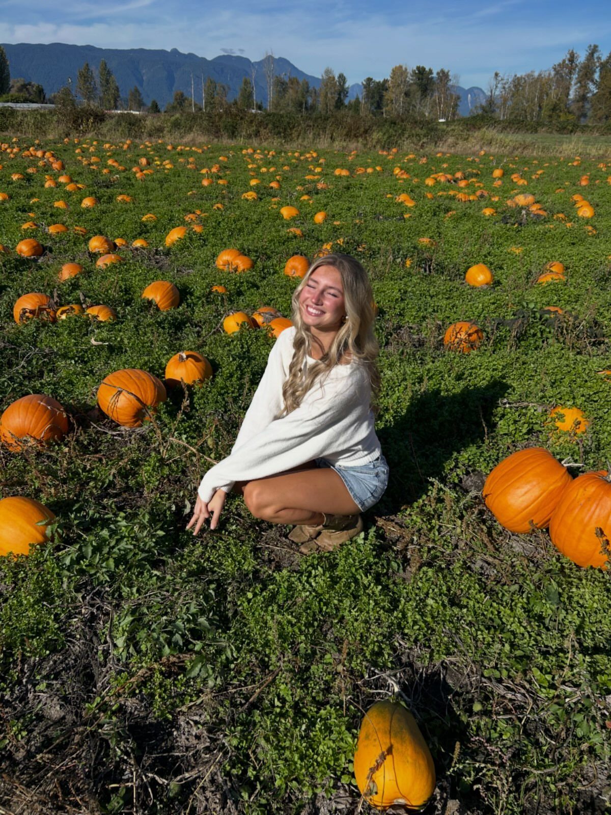 Woman in white sweater and denim shorts crouching in a pumpkin patch field.