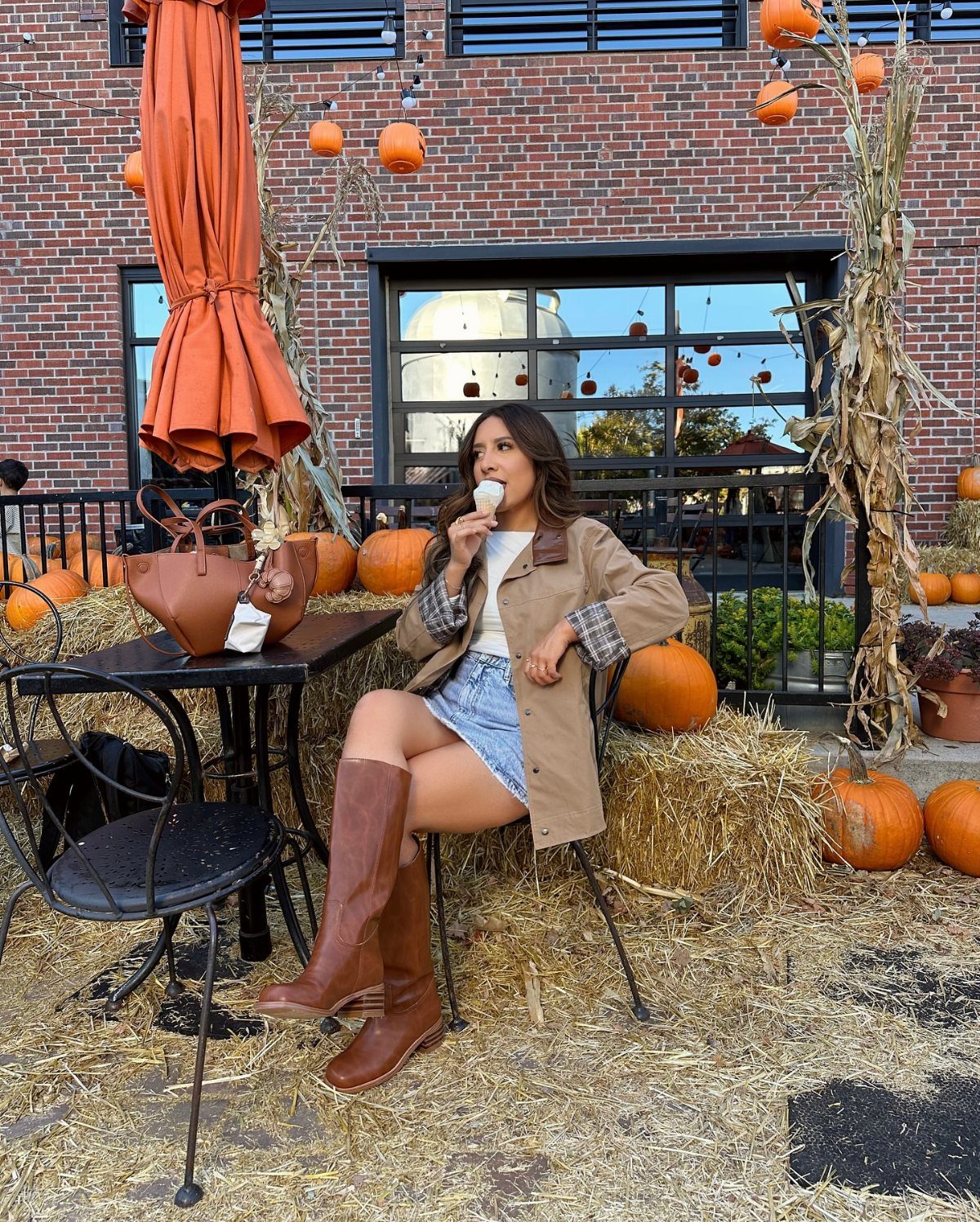 Woman in tan shacket and brown boots sitting on hay with pumpkins.