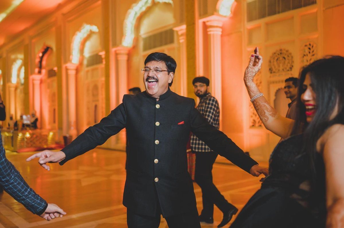 Man in black bandhgala suit dancing in an ornate, warmly lit hall.