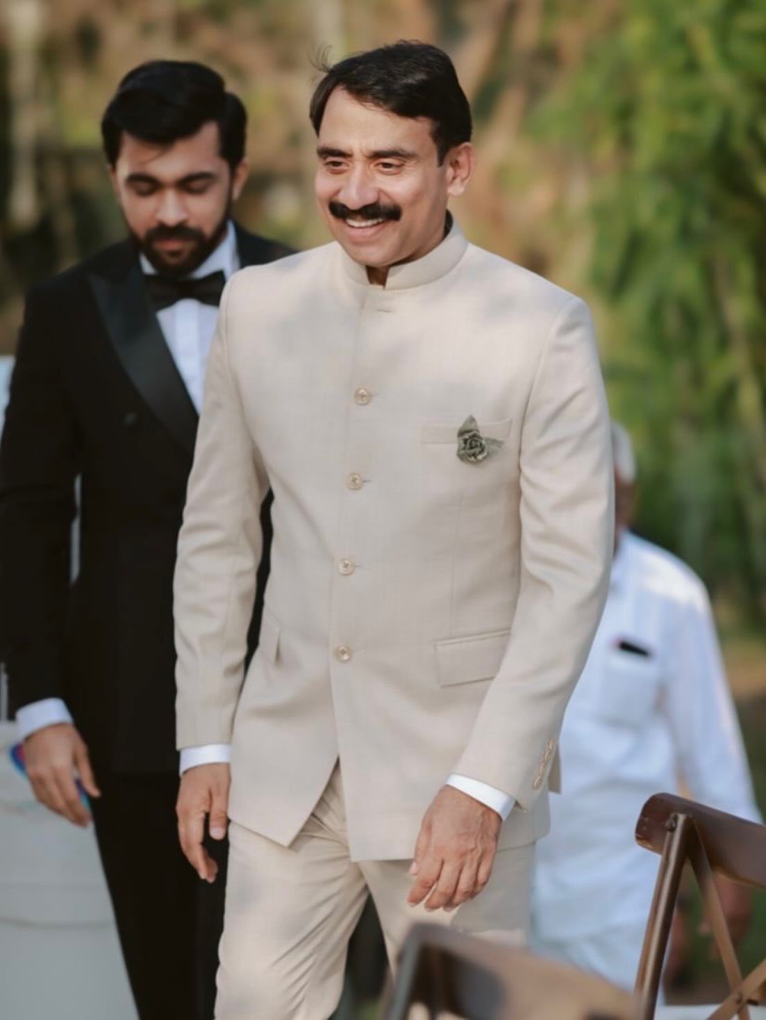 Man in cream Nehru suit smiling next to man in black tuxedo outdoors