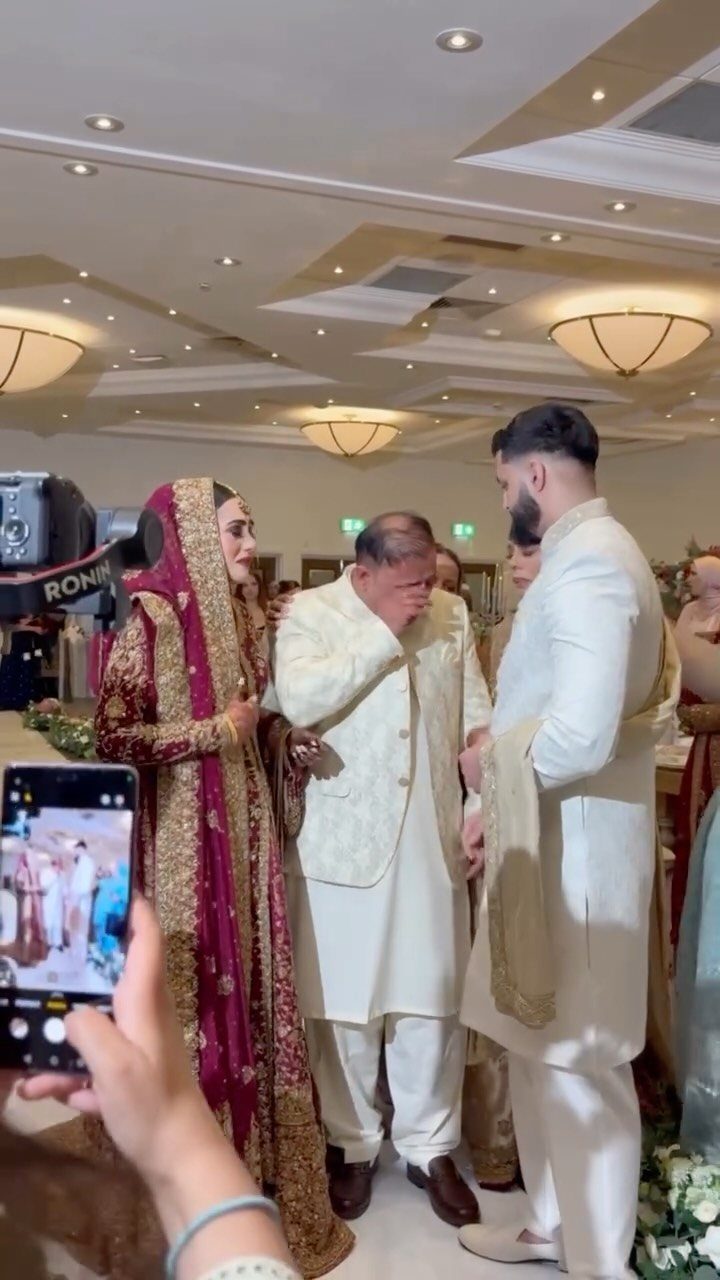Man in cream traditional attire standing with a bride and groom indoors.