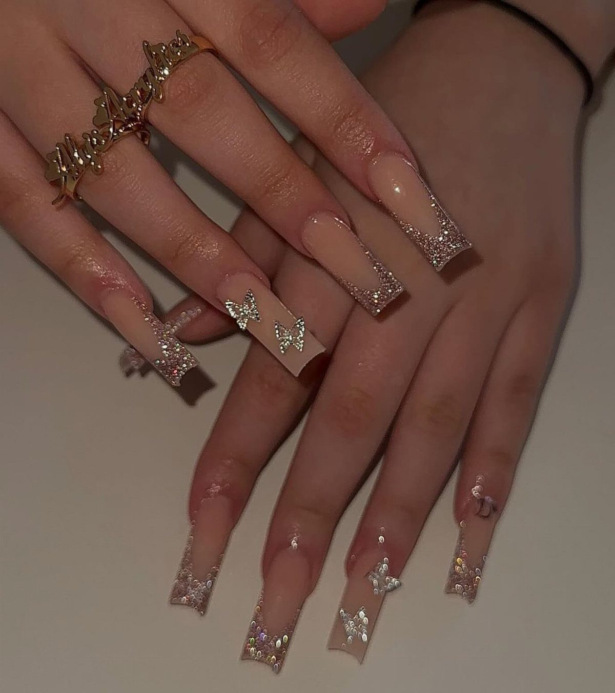 Long square nails with nude base, silver glitter French tips and butterfly charms.