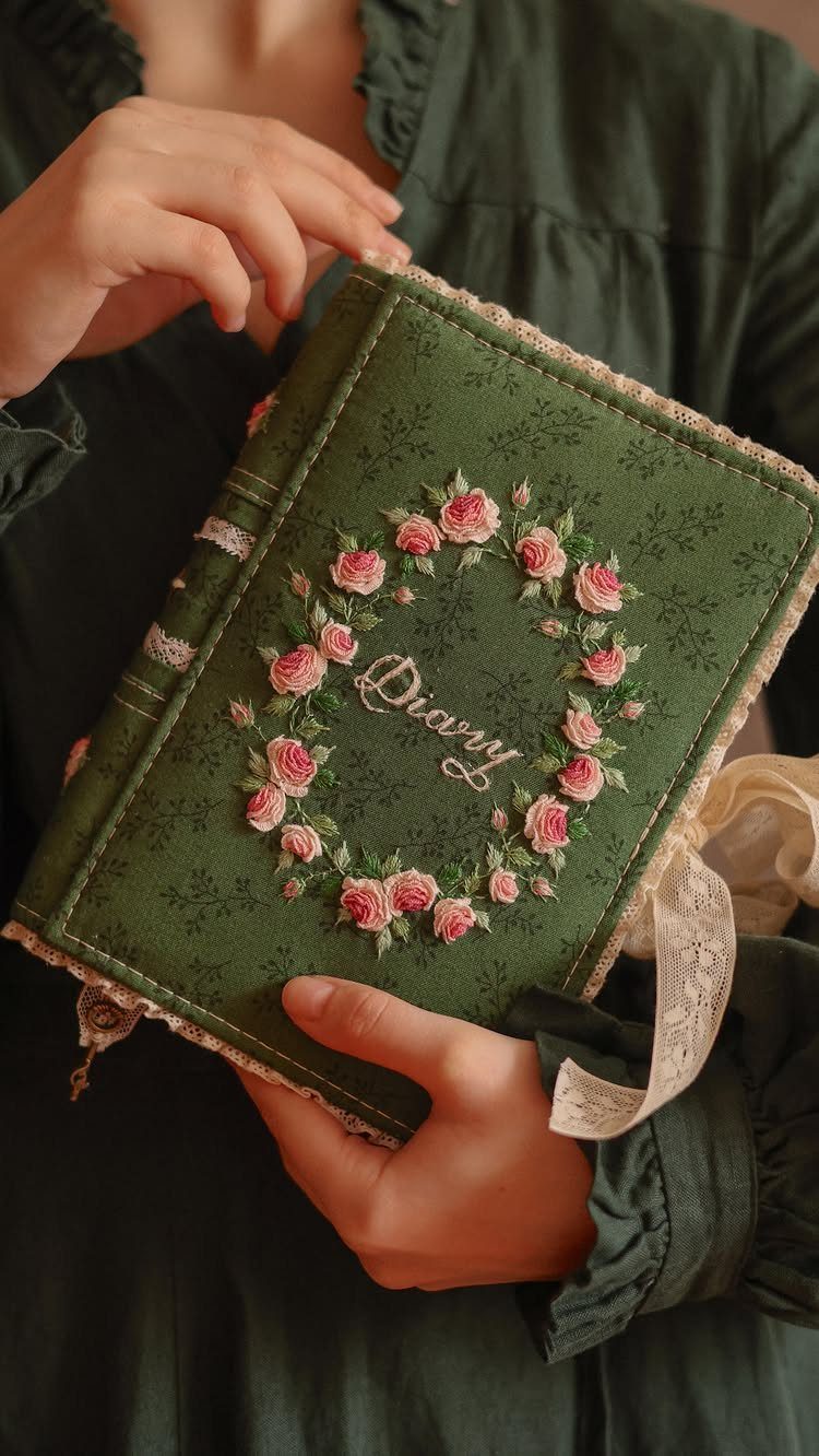 Green fabric diary with a pink rose wreath and lace trim held by hands.