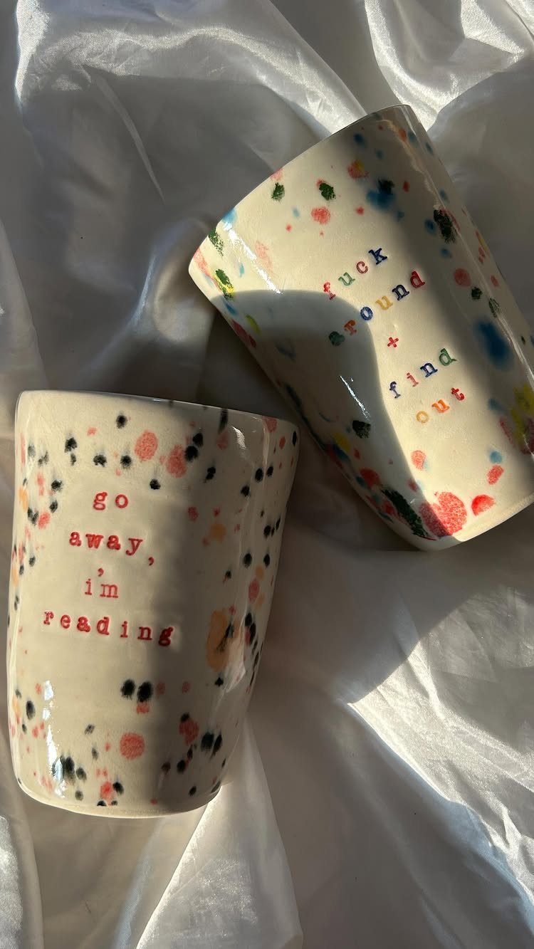 Two speckled ceramic mugs with red text resting on white wrinkled fabric.