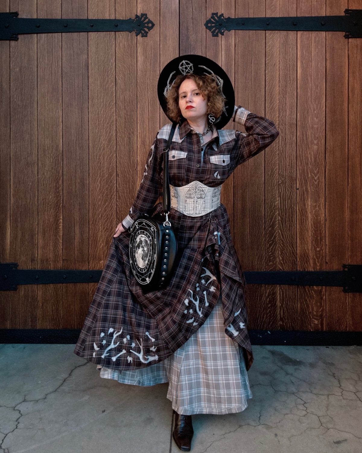 Woman in brown plaid dress and black witch hat posing by wooden doors.