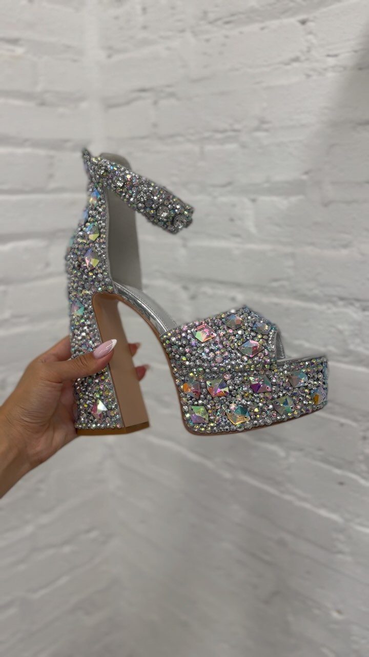 Silver platform high-heeled sandal covered in sparkling iridescent rhinestones held by a hand.