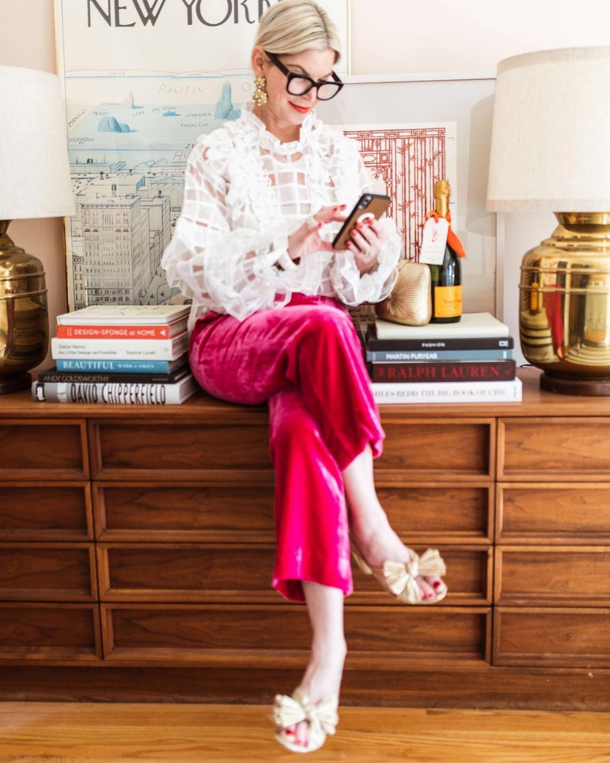 Woman in white lace blouse and pink satin pants sitting on wooden dresser.