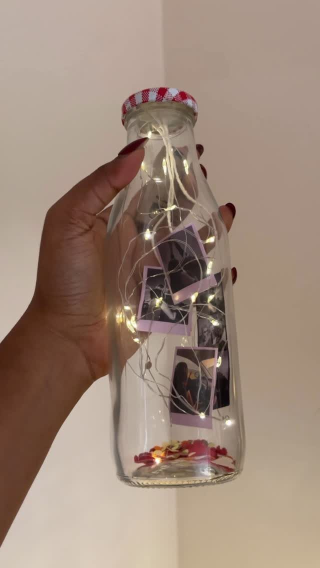 Glass bottle containing polaroid photos, fairy lights, and red petals held by a hand.