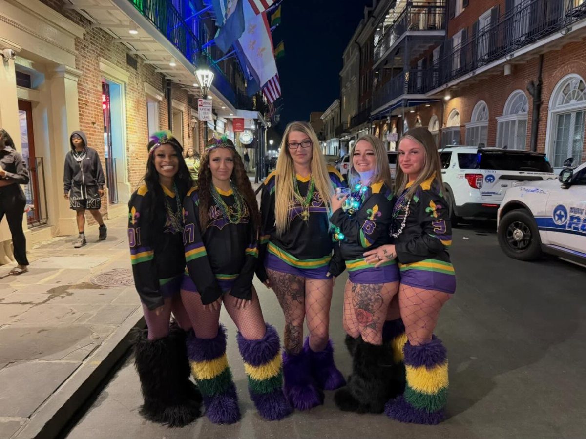 Five women in purple, green, and gold Mardi Gras outfits posing on a street.