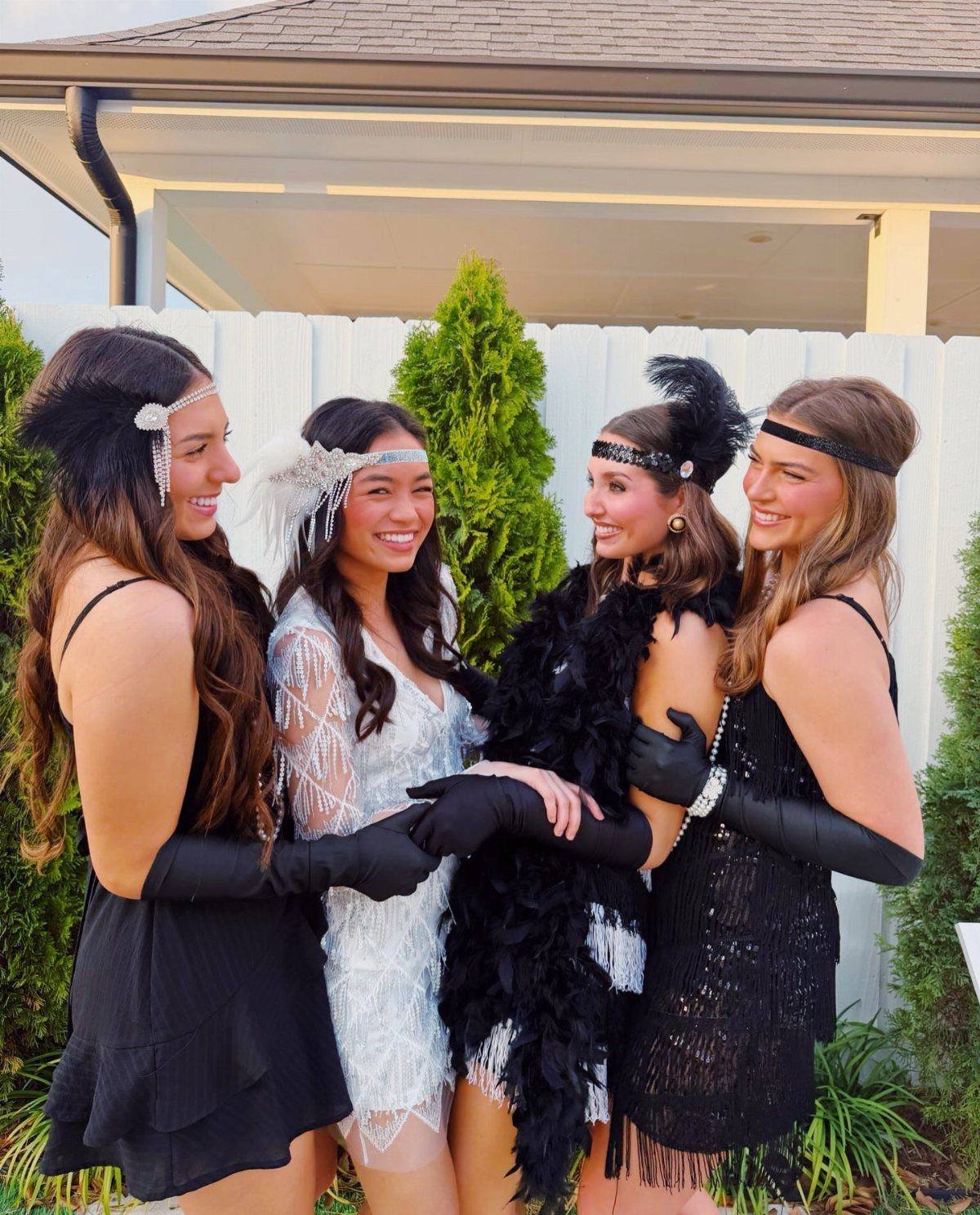 Four women wearing 1920s flapper dresses, headbands, and long black gloves outdoors.