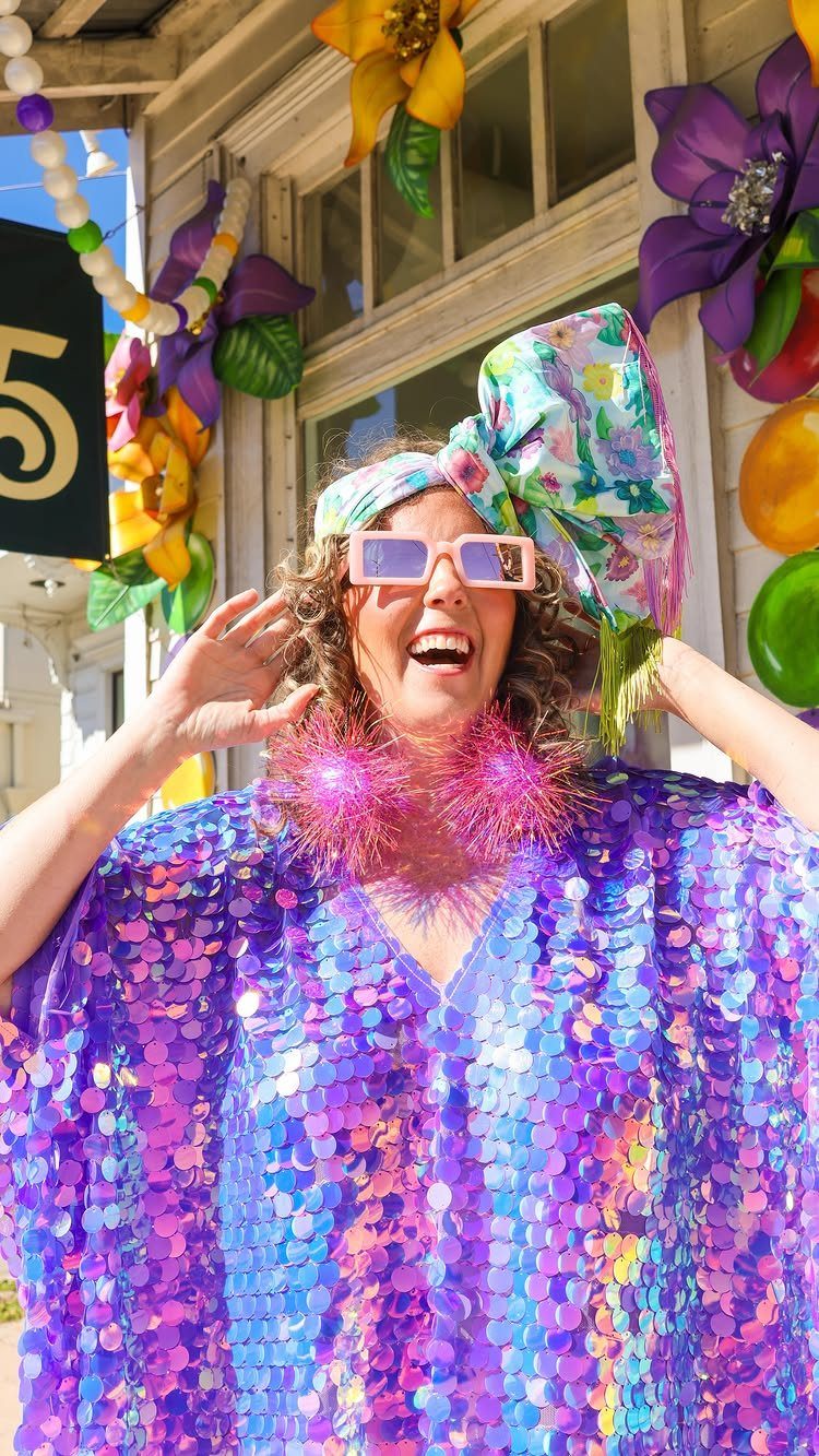 Woman in a purple sequined outfit with a floral headwrap and pink sunglasses.