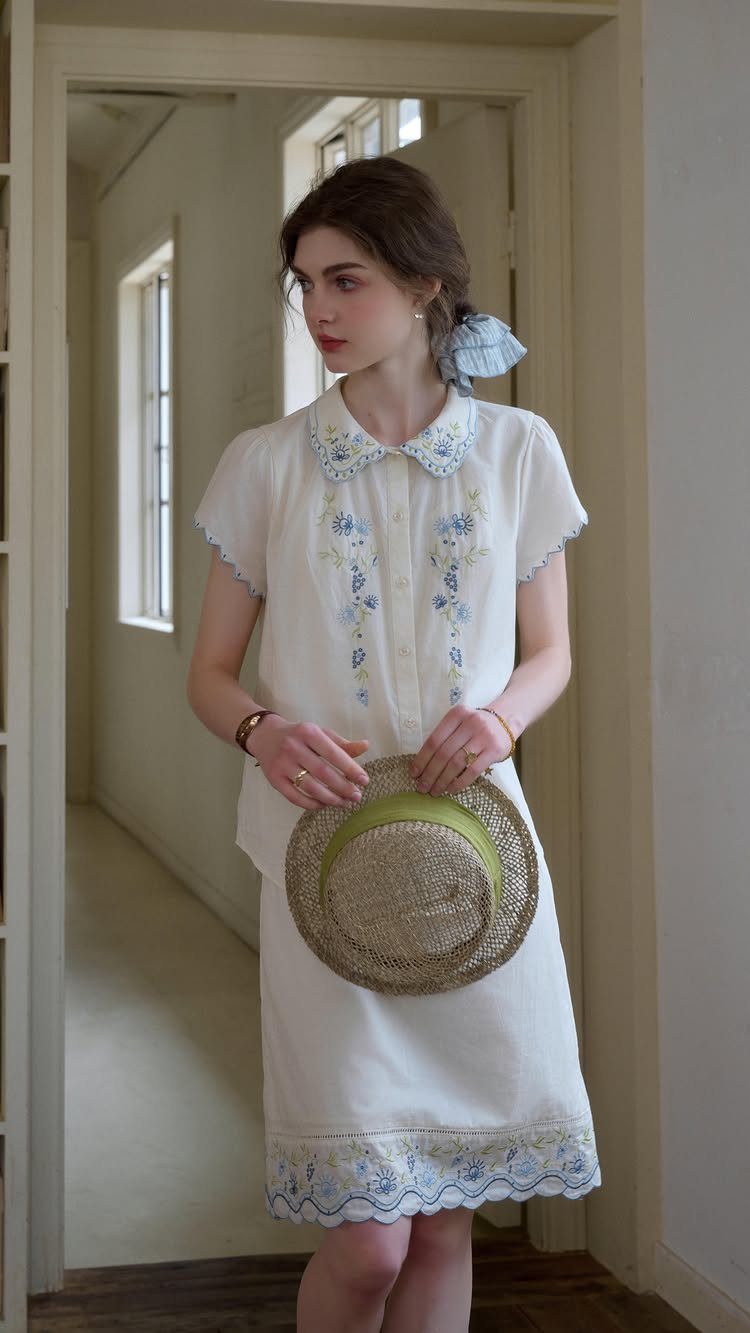 Woman in a white dress with blue floral embroidery holding a straw hat.