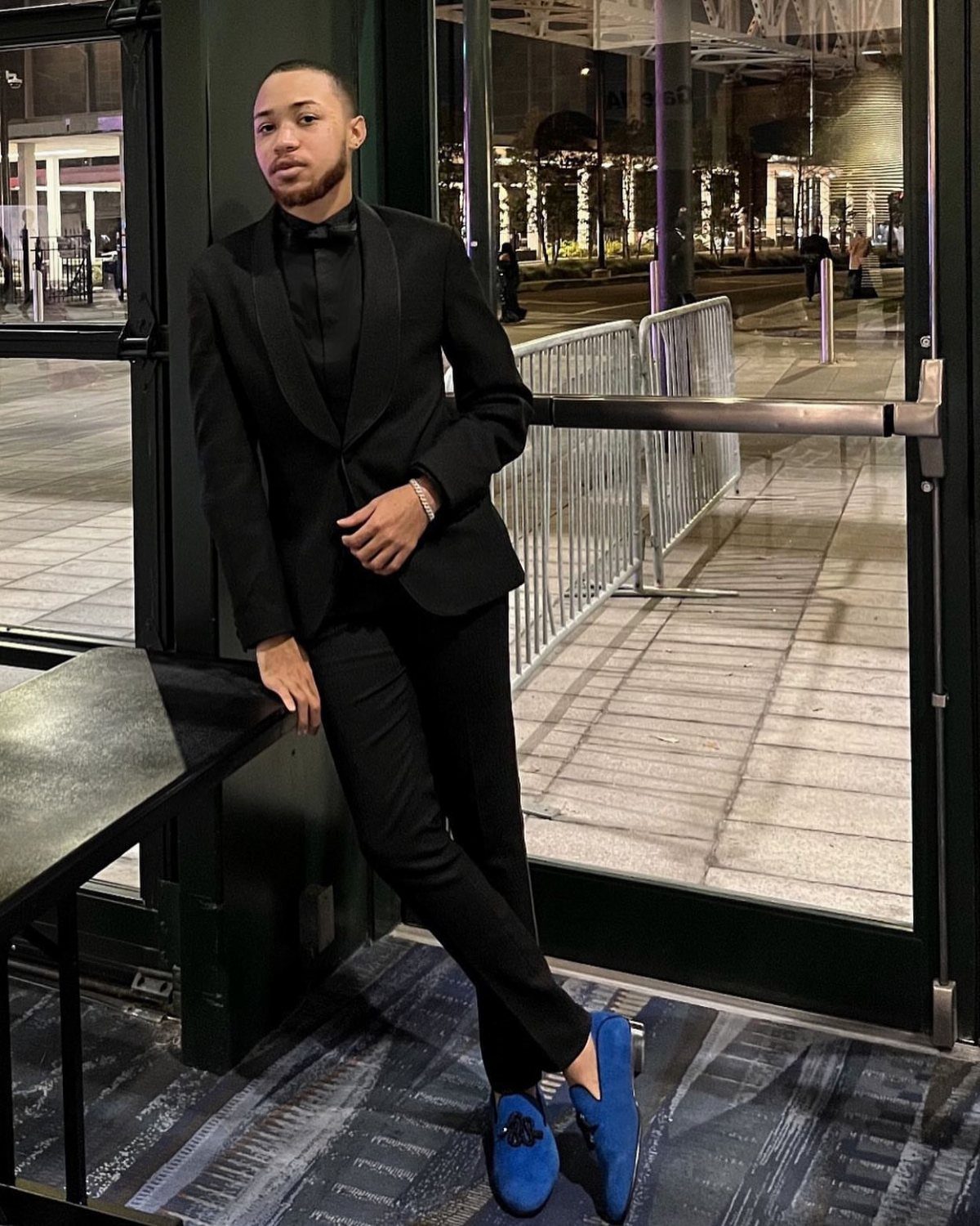 Man in a black tuxedo and blue velvet loafers posing by a glass door.
