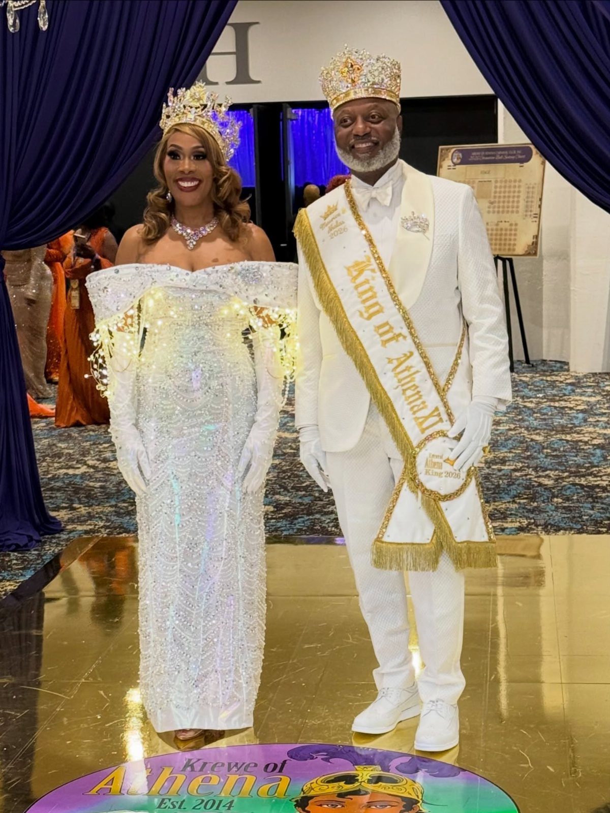 Man and woman in formal white Mardi Gras ball outfits with crowns and sash.