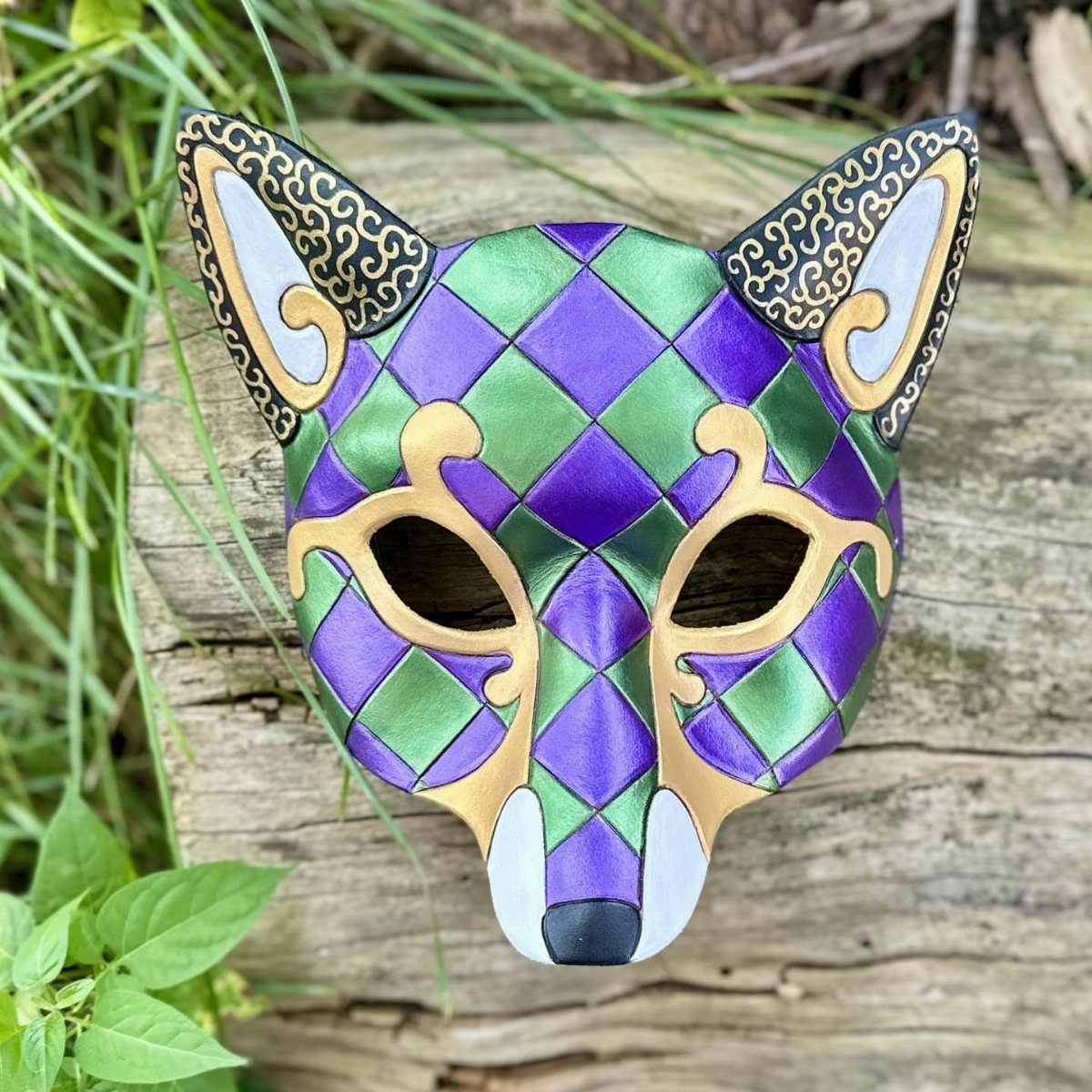 Fox-shaped masquerade mask with purple and green diamond patterns on a wooden log
