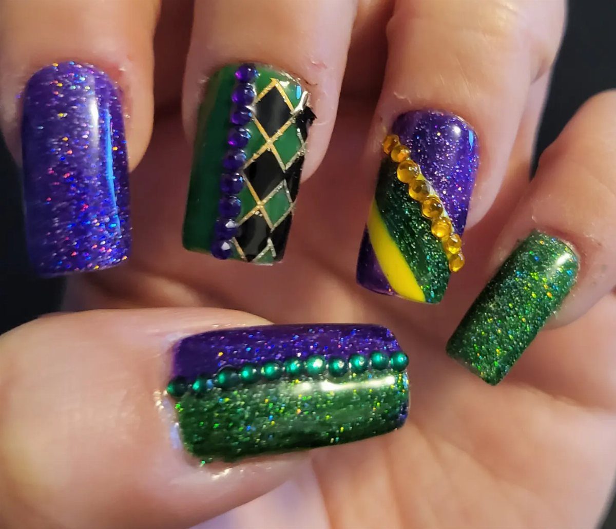 Short square nails with purple, green, and gold glitter and decorative bead accents.
