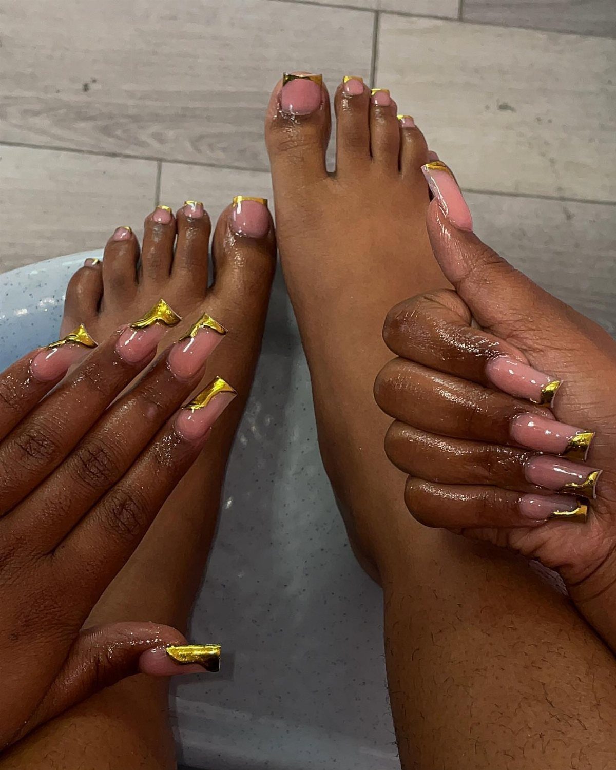 Hands and feet with nude nails and metallic gold French tips on tiles.