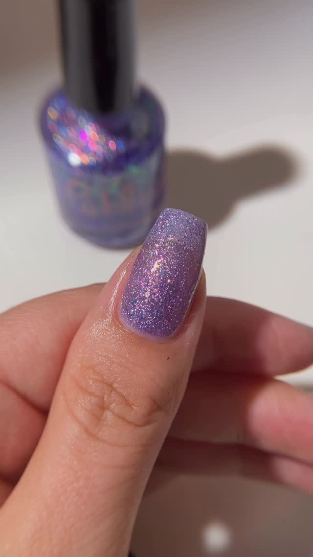 Holographic purple glitter nail polish on a thumb with polish bottle behind.