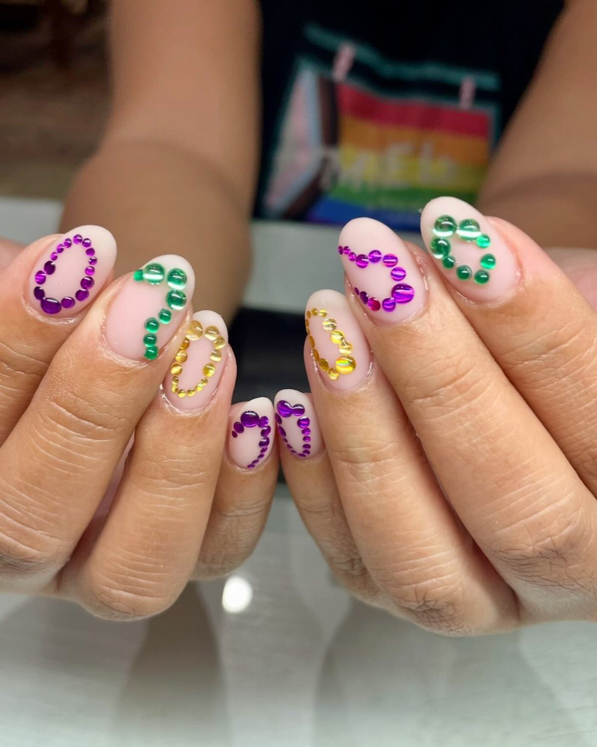 Manicured nails with purple, green, and gold rhinestone circles on pink polish.