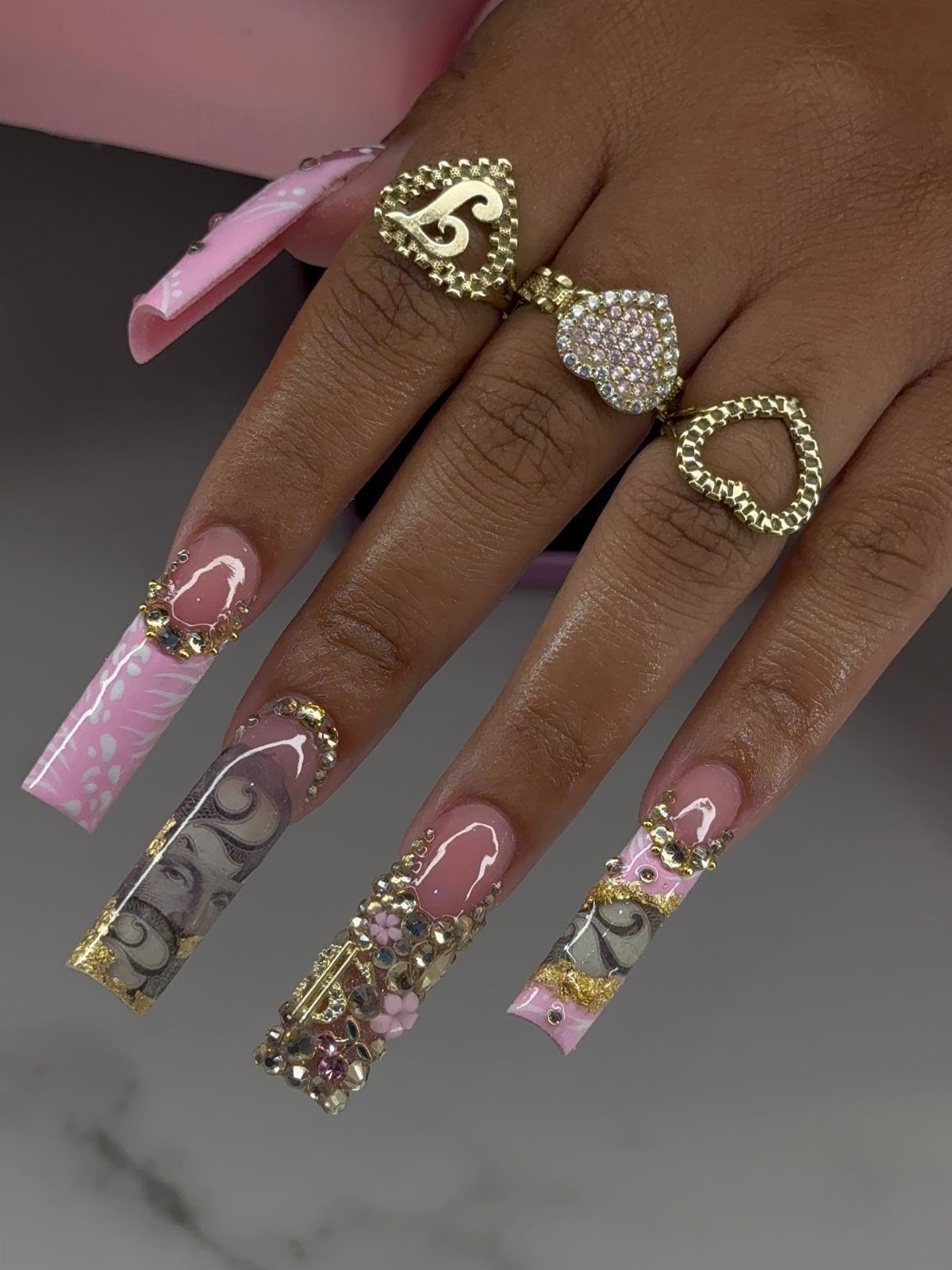 Long acrylic nails decorated with rhinestones, jewels, and pink and grey patterns.