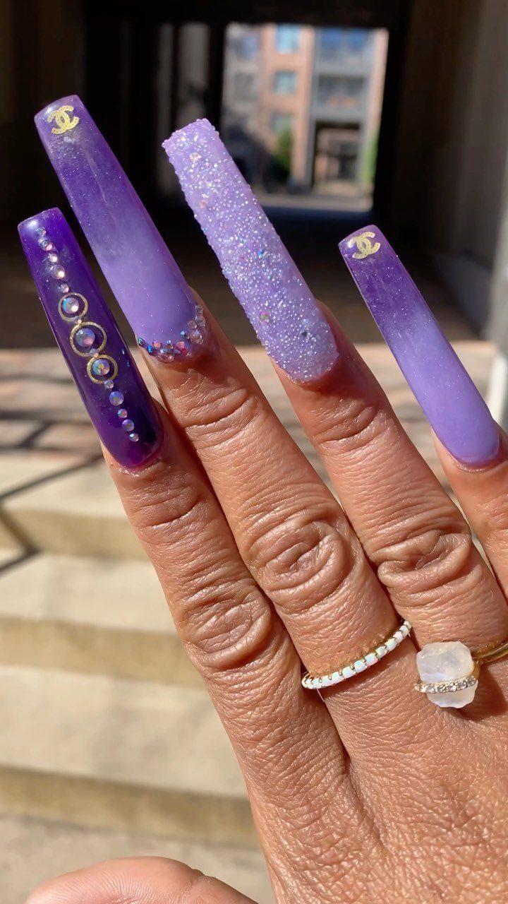 Long purple ombre and glitter nails with gold logos and small rhinestones.