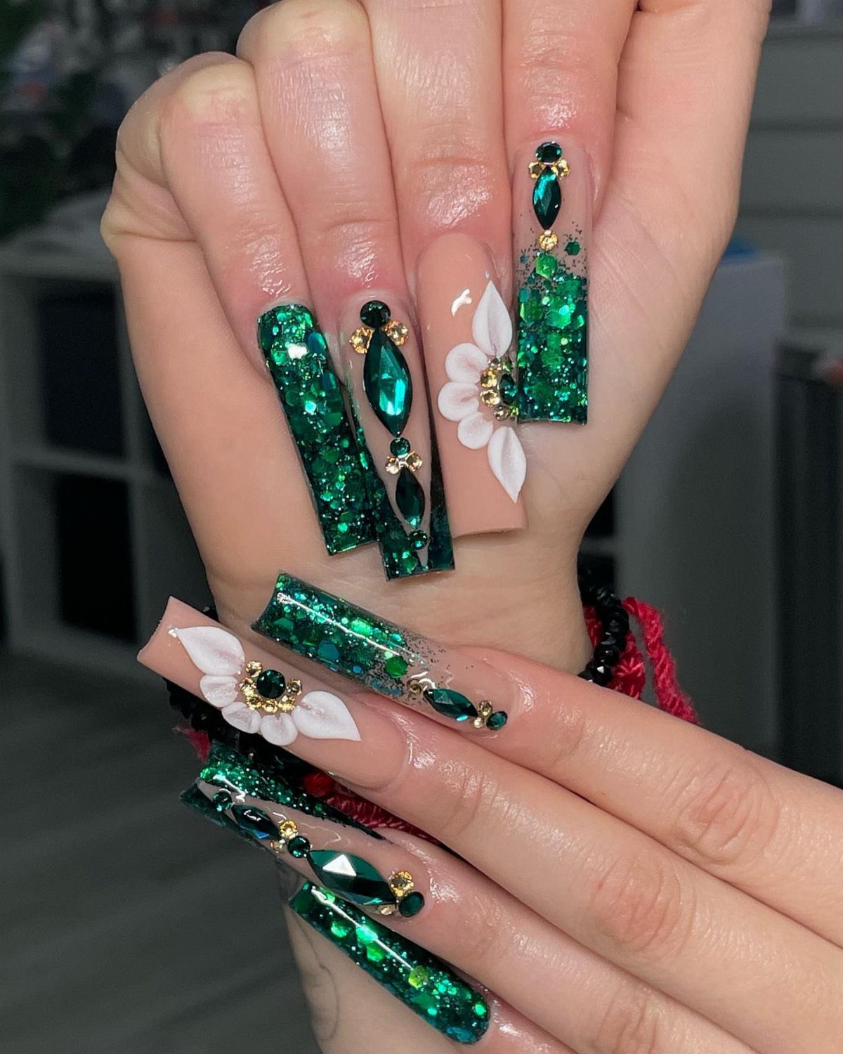 Long green glitter nails decorated with white 3D flowers and emerald gemstones.